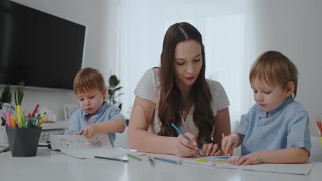 A-young-mother-with-two-children-sitting-at-a-white-table-draws-colored-pencils-on-paper-in-slow-motion